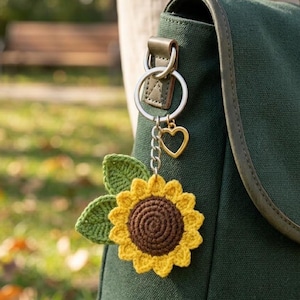 May include: A crochet sunflower keyring featuring a dark brown centre with bright yellow petals and two green leaves. The sunflower is attached to a silver metal split ring with a small gold-toned heart charm hanging from the chain link.