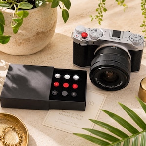 Op de afbeelding: Een zilverkleurige Fujifilm X-M5 camera met een zwarte lens en een rode ontspanknop. Daarnaast staat een zwarte doos met negen ronde ontspanknoppen in zilver, rood en zwart, met vlakke, holle en bolle oppervlakken.