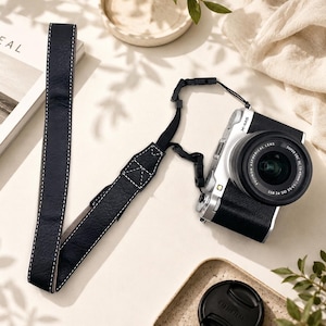 Może przedstawiać: Czarny pasek do aparatu z białymi kontrastowymi przeszyciami przymocowany do srebrno-czarnego bezlusterkowca Fujifilm X-M5 z obiektywem. Pasek ma teksturowane wykończenie i jest połączony z korpusem aparatu za pomocą czarnych nylonowych zaczepów.