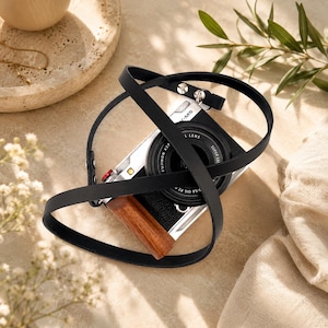 May include: A silver digital camera with a black lens and a brown wooden grip, featuring a long, thin black leather strap that loops around the camera body. The camera has a small red button on the top and the text X-M5 printed on the front.