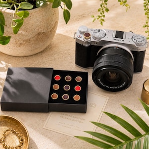 Könnte beinhalten: Eine silberne Fujifilm X-M5 Kamera mit schwarzem Objektiv steht neben einer schwarzen Schachtel mit neun runden Kamera-Auslöserknöpfen in Rot, Braun und Violett. Eine kleine Karte trägt die Aufschrift CAMERA SHUTTER RELEASE BUTTON, BLACK, SILVER, RED, FLAT, CONCAVE, CONVEX.