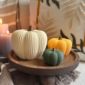 Pode incluir: Três abóboras decorativas caneladas nas cores creme, amarelo e verde escuro, cada uma com um pequeno caule castanho no topo. As abóboras estão dispostas num tabuleiro redondo de madeira castanha escura com uma base central única.
