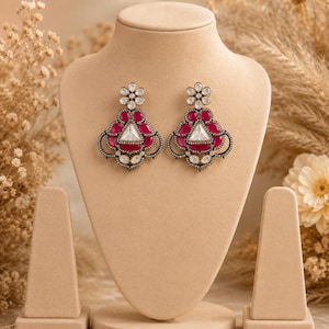 Puede incluir: Un par de pendientes colgantes ornamentados con una piedra central triangular transparente rodeada de piedras de color rosa intenso en forma de lágrima. El diseño incluye un poste superior en forma de flor y detalles de metal oscuro incrustados con pequeños cristales.