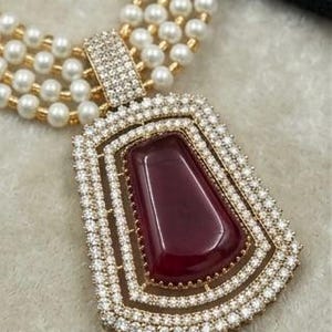 May include: A multi-strand pearl necklace with a large pendant. The pendant features a rectangular, deep red gemstone surrounded by a gold frame and encrusted with small, clear crystals. The necklace has a gold clasp.