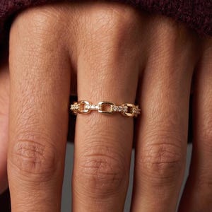 May include: A gold-toned ring featuring a repeating chain-link design. The surface of the metal links is embellished with small, clear, sparkling stones set along the top, creating a continuous, textured band that encircles the finger.