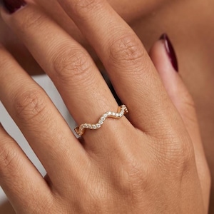 May include: A gold ring featuring a wavy, serpentine band design. The surface of the band is set with a continuous row of small, clear, round-cut stones that follow the undulating shape of the metal, creating a delicate and shimmering appearance.