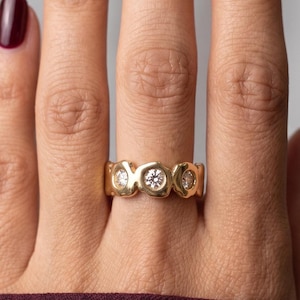 May include: A gold ring featuring three round, clear, faceted stones set in a continuous, organic, polished metal band. The band has a smooth, rounded, and slightly irregular shape that encircles the finger, showcasing a warm, reflective finish.
