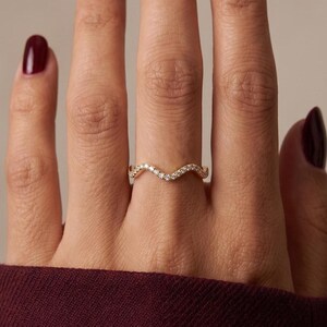 May include: A gold ring featuring a delicate, wavy band design set with a series of small, clear, sparkling stones along the top curve. The ring is worn on a finger, showcasing the thin, metallic band and the embedded stones in a chevron shape.