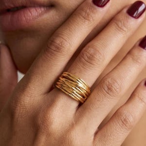 May include: A wide gold ring featuring a textured design composed of multiple thin, overlapping, and intertwined metallic bands that create a layered, wire-like appearance across the surface of the jewelry piece.