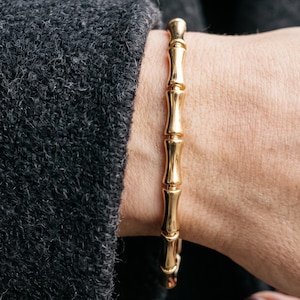 May include: A polished gold bracelet featuring a series of connected, cylindrical segments that resemble the natural texture and nodes of bamboo stalks. The metallic finish is smooth and reflective, creating a continuous, uniform design.