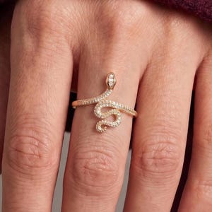 May include: A gold-toned ring shaped like a coiled snake, featuring a textured surface embedded with numerous small, clear, sparkling stones along the entire length of the body and a single, slightly larger clear stone set at the head.