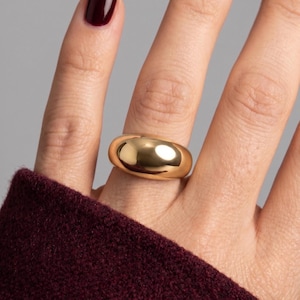 Puede incluir: Un anillo de tono dorado pulido con una banda gruesa, lisa y redondeada en forma de cúpula. La superficie del anillo es altamente reflectante, mostrando un brillo intenso de la fuente de luz, creando un aspecto metálico sólido.