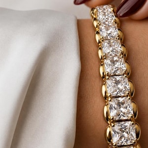 May include: A gold-toned tennis bracelet featuring a continuous row of square-cut, clear, faceted stones set in polished metal links. The bracelet is worn on a wrist, positioned next to a draped, smooth, off-white fabric with a soft sheen.