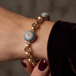 Può includere: Un bracciale a maglie dorate con ciondoli circolari incastonati con pietre trasparenti, turchesi e blu scuro. Il ciondolo centrale presenta un motivo concentrico di queste pietre, creando un dettaglio simile a un occhio lungo le maglie.