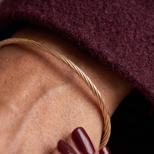 Puede incluir: Una pulsera tipo brazalete fina de color dorado con una textura trenzada similar a una cuerda. La banda metálica tiene un acabado pulido que refleja la luz a lo largo de su superficie en espiral, creando un patrón uniforme y continuo.
