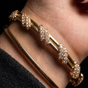 Puede incluir: Un brazalete dorado que presenta una serie de acentos diagonales de piedras claras brillantes envueltas alrededor de la banda, junto con un segundo brazalete dorado liso y sencillo, usados juntos en la muñeca.