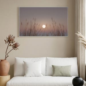 May include: A framed landscape print featuring a sunset over tall grass. The sun is a bright white orb in a dusky pink and purple sky. The print hangs above a white sofa with decorative pillows and a black vase.