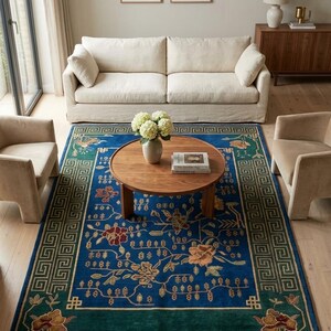 May include: A rectangular area rug featuring a vibrant blue center with floral patterns in red, gold, and green. The rug has a dark green border with a geometric Greek key design and fringe on the ends, placed under a round wooden coffee table.