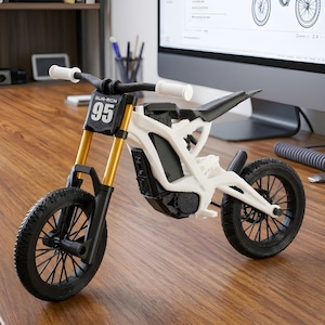 Może przedstawiać: Biało-czarny miniaturowy motocykl ze złotymi przednimi widelcami. Kierownica ma białe uchwyty, a przód motocykla wyświetla tekst "SUR-RON" nad numerem "95". Opony są czarne, a motocykl stoi na drewnianej powierzchni.