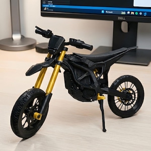 Może przedstawiać: Czarny i złoty miniaturowy model motocykla. Model posiada czarną ramę, koła i siedzenie, ze złotymi przednimi widelcami i akcentami. Opony mają szczegółowy wzór bieżnika. Motocykl jest wystawiony na jasnej powierzchni.