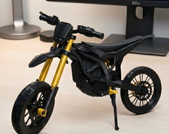 May include: A black and gold miniature motorcycle model. The model features a black frame, wheels, and seat, with gold-colored front forks and accents. The tires have a detailed tread pattern. The motorcycle is displayed on a light-colored surface.