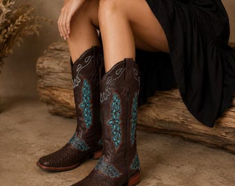 May include: Dark brown leather cowboy boots featuring a textured finish and intricate turquoise embroidery along the shaft and foot. The boots have a square toe, a low stacked heel, and pull tabs at the top of the shaft for easy wear.