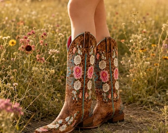 Puede incluir: Un par de botas vaqueras de cuero marrón con intrincados bordados florales en tonos rosa, blanco y azul. Las botas tienen punta cuadrada, tacón bajo de bloque y ribetes de color turquesa brillante a lo largo de las costuras laterales.