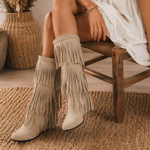 May include: A pair of knee-high beige boots featuring three distinct tiers of long, flowing fringe that cover the shaft. The boots have a rounded toe, a flat sole, and a soft, matte finish, extending from the ankle up to just below the knee.