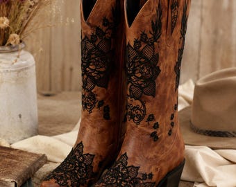 May include: A pair of mid-calf brown leather boots featuring intricate black lace floral overlays on the toe and shaft. The boots have a pointed toe, a stacked block heel, and a scalloped top edge with pull tabs on the sides of each boot.