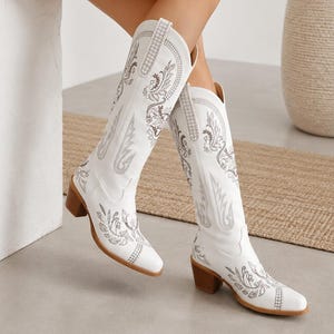 May include: A pair of white knee-high boots featuring intricate silver embroidery in floral and paisley patterns. The boots have a pointed toe, a stacked block heel, and pull tabs at the top of the shaft. The design includes detailed stitching.
