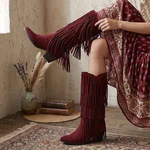 May include: A pair of knee-high boots in a deep burgundy colour, featuring multiple tiers of long fringe detailing along the shaft and a block heel. The boots are crafted with a soft, suede-like texture and have a pointed toe design.