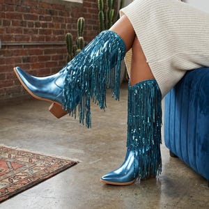 May include: A pair of metallic blue knee-high boots featuring a pointed toe, a stacked wooden block heel, and long, shimmering fringe detailing that covers the entire shaft of the boot, creating a dynamic and textured appearance.
