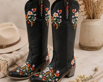 May include: A pair of black knee-high boots featuring colorful floral embroidery on the toes and shafts. The boots have a pointed toe, a stacked block heel, and pull tabs at the top. The embroidery includes red, orange, white, and green floral patterns.