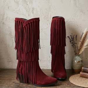 May include: A pair of deep burgundy suede boots featuring three distinct tiers of long, flowing fringe that extend from the top down to the ankle. The boots have a pointed toe, a sturdy block heel, and a dark interior lining visible at the opening.
