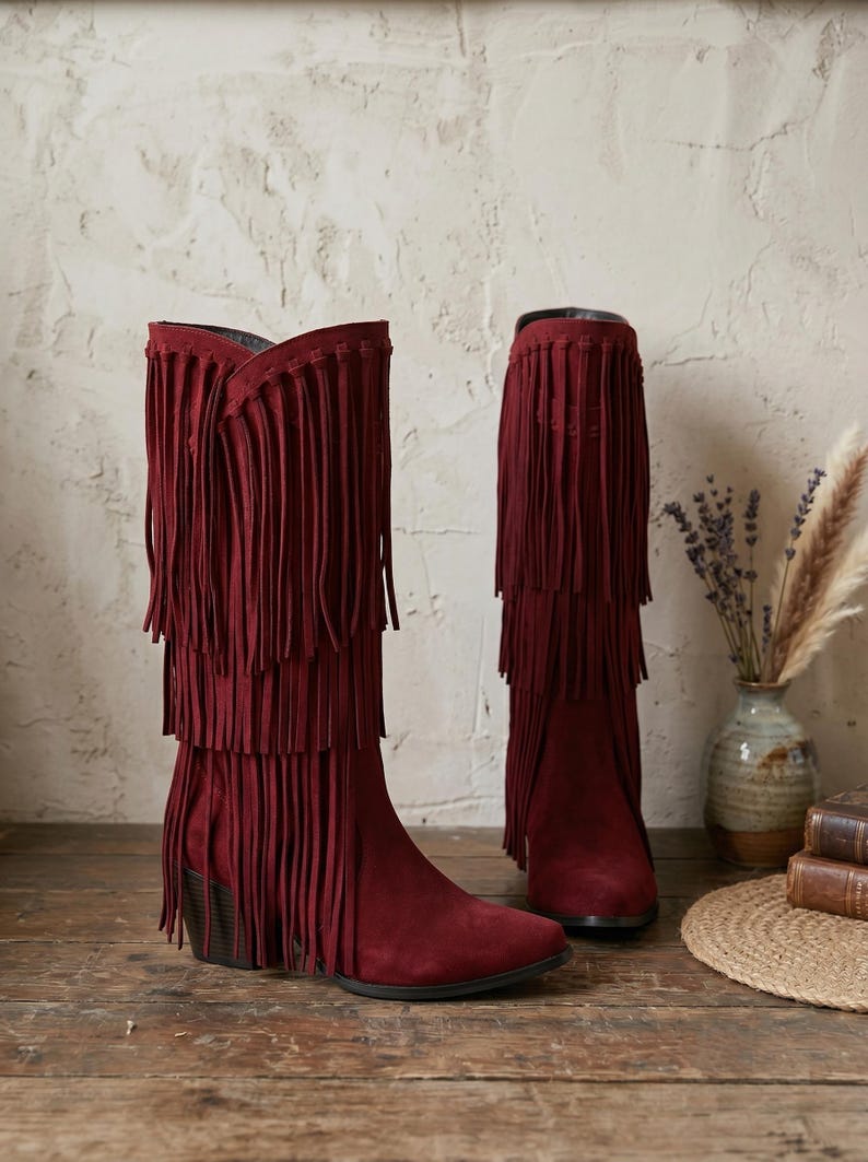 May include: A pair of deep burgundy suede boots featuring three distinct tiers of long, flowing fringe that extend from the top down to the ankle. The boots have a pointed toe, a sturdy block heel, and a dark interior lining visible at the opening.
