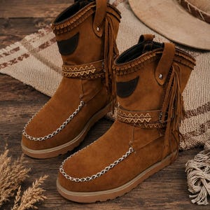 Könnte beinhalten: Ein Paar wadenhohe Stiefel aus braunem Wildleder mit Fransen, einem dekorativen Riemen mit geometrischem Muster und weißer Ziernaht entlang der abgerundeten Schuhspitze. Die Stiefel haben eine flache Gummisohle und einen dunkelbraunen Aufnäher.