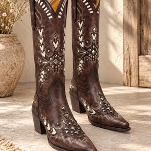 May include: A pair of dark brown leather cowboy boots featuring a pointed toe, a stacked heel, and intricate white stitching. The boots are decorated with silver-toned metal studs and geometric accents arranged in patterns along the shaft and toe.