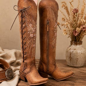 May include: A pair of knee-high brown leather boots featuring intricate floral embroidery in white and pink thread. The left boot has a front lace-up closure with dark brown laces, while the right boot features a side zipper and a pointed toe.