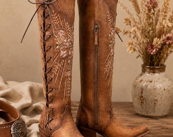 May include: A pair of knee-high brown leather boots featuring intricate floral embroidery in white and pink thread. The left boot has a front lace-up closure with dark brown laces, while the right boot features a side zipper and a pointed toe.