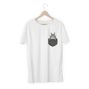 Pu&ograve; includere: T-shirt bianca con tasca con illustrazione a cartoni animati di Totoro. La tasca &egrave; grigio scuro e il personaggio di Totoro &egrave; grigio con la pancia color crema. La maglietta &egrave; appesa a una gruccia di legno.