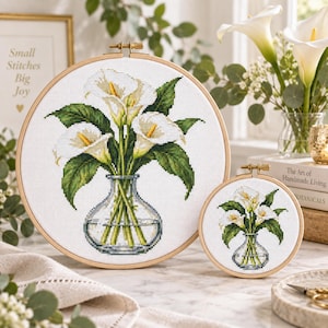 May include: Two circular wooden embroidery hoops containing cross-stitch fabric. The fabric features a design of white calla lilies with green leaves arranged in a clear glass vase. The larger hoop is on the left and the smaller hoop is on the right.
