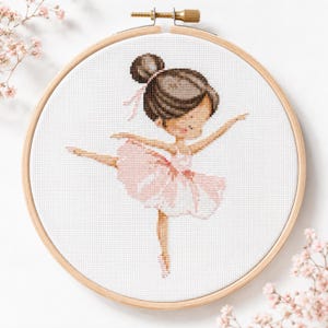 Puede incluir: Un bordado de punto de cruz en un bastidor de madera redondo que muestra a una bailarina de pelo castaño con un tutú rosa. La bailarina aparece en una pose elegante con los brazos extendidos y una pierna levantada sobre tela blanca cuadriculada.