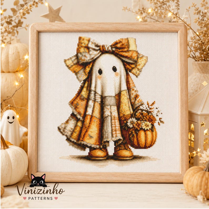 May include: A cross-stitch pattern in a light wood frame featuring a ghost wearing a large plaid bow and a plaid cape, holding a small pumpkin filled with flowers. The ghost has two black eyes and is rendered in shades of orange, brown, and white.