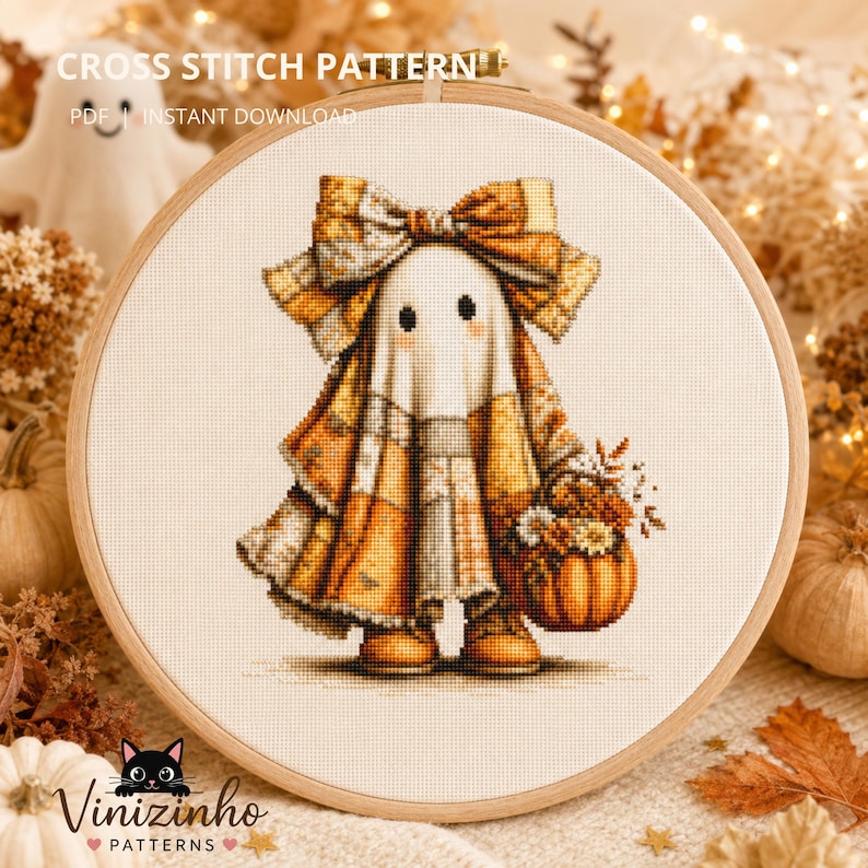 Cross stitch hoop featuring a cute patchwork ghost wearing a large autumn bow and holding a pumpkin basket. Stitched on cream 14-count Aida fabric with warm orange, beige, and brown tones, displayed in a wooden embroidery hoop surrounded by cozy Halloween fall decor, pumpkins, dried flowers, and soft fairy lights. Cottagecore Halloween embroidery pattern.