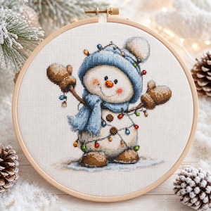Peut inclure: Une broderie au point de croix dans un tambour en bois rond représentant un bonhomme de neige portant un bonnet et une écharpe en tricot bleu, des moufles et des bottes marron. Il est entouré d'une guirlande lumineuse colorée sur fond blanc.