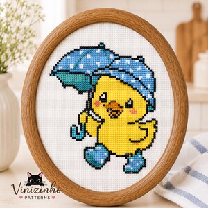 May include: Cross-stitch art of a yellow chick wearing blue rain boots and holding a blue umbrella with white polka dots. The artwork is framed in a round wooden frame. The brand name "Vinizinho Patterns" is at the bottom.