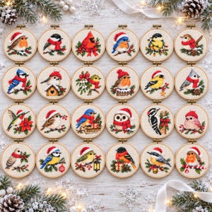 Op de afbeelding: Een verzameling van twintig ronde houten borduurringen, elk met een kruissteekontwerp van een vogel met winteraccessoires zoals een muts of sjaal, omringd door feestelijke elementen zoals hulst, dennentakken en kerstversieringen.