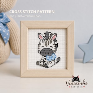 Op de afbeelding: Een ingelijst kruissteekpatroon met een cartoon zebra met een blauwe strik. De zebra is zwart, wit en grijs, met roze accenten. De lijst is van licht hout. De tekst "CROSS STITCH PATTERN" is zichtbaar.