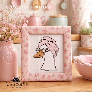 Op de afbeelding: Een kruissteekpatroon met een witte gans die een roze handdoek op het hoofd en een bril met zwart montuur draagt. Het ontwerp wordt getoond in een vierkant frame met een roze bloemenstoffen rand, tegen een lichte keukenachtergrond.