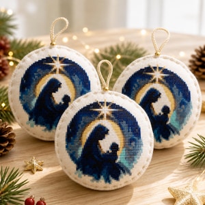 May include: Three round, cream-colored fabric ornaments featuring a cross-stitched nativity scene. Each ornament displays a dark blue silhouette of a figure holding a child against a golden crescent moon and a bright star, finished with a gold hanging loop.